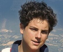 Retrato do Beato Carlo Acutis sorrindo, usando uma camisa polo vermelha e uma mochila, com o Santuário de Assis ao fundo